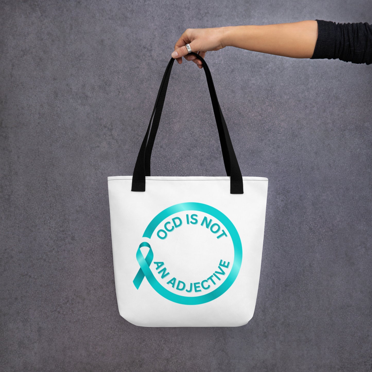 A hand holds NeuroNiche Design’s "OCD Is Not An Adjective" white tote bag with black straps, a teal ribbon, and the slogan in teal on the front. The background is plain gray.