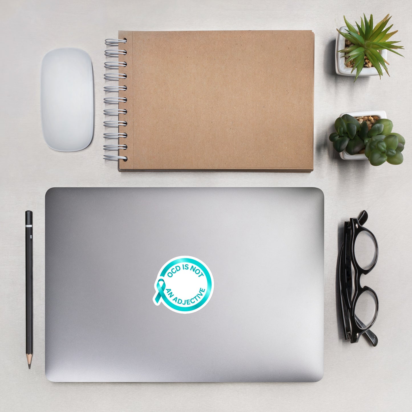 A laptop displays the NeuroNiche Design "OCD is Not An Adjective" sticker—its durable vinyl ensures a bubble-free, long-lasting style—next to a notebook, wireless mouse, pencil, glasses, and two small potted plants.