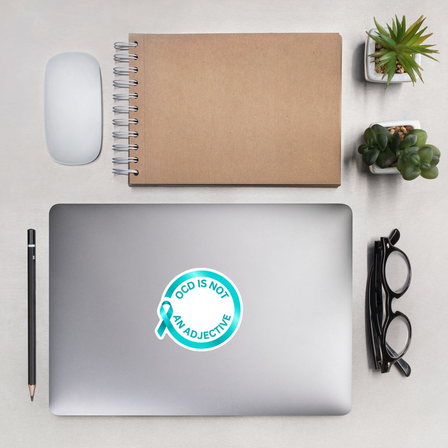 A laptop with a durable NeuroNiche Design "OCD is Not An Adjective" vinyl sticker sits on a desk, surrounded by a notebook, mouse, pencil, eyeglasses, and two neatly arranged small potted plants.