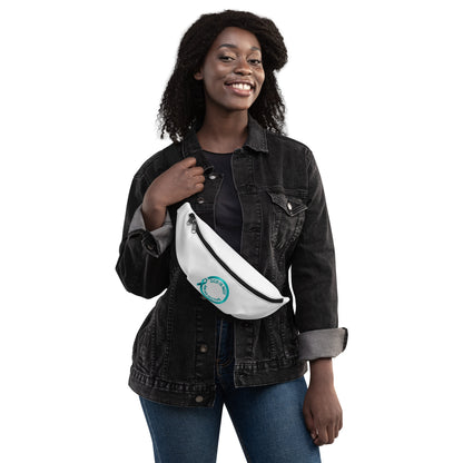 A smiling woman in a black denim jacket and blue jeans stands holding the "OCD Is Not An Adjective" white fanny pack by NeuroNiche Design, featuring a turquoise circular logo and adjustable straps, slung across her torso against a white background.