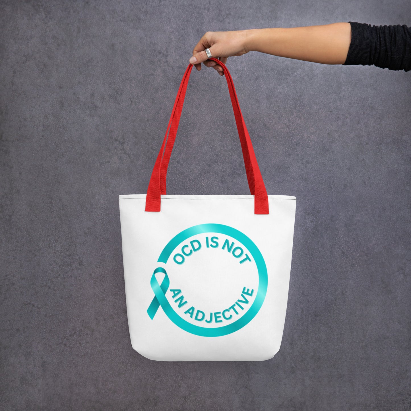 A hand holds the "OCD Is Not An Adjective" tote by NeuroNiche Design, a spacious white bag with red handles, featuring a teal ribbon and a circle with the phrase "OCD IS NOT AN ADJECTIVE," set against a gray background.