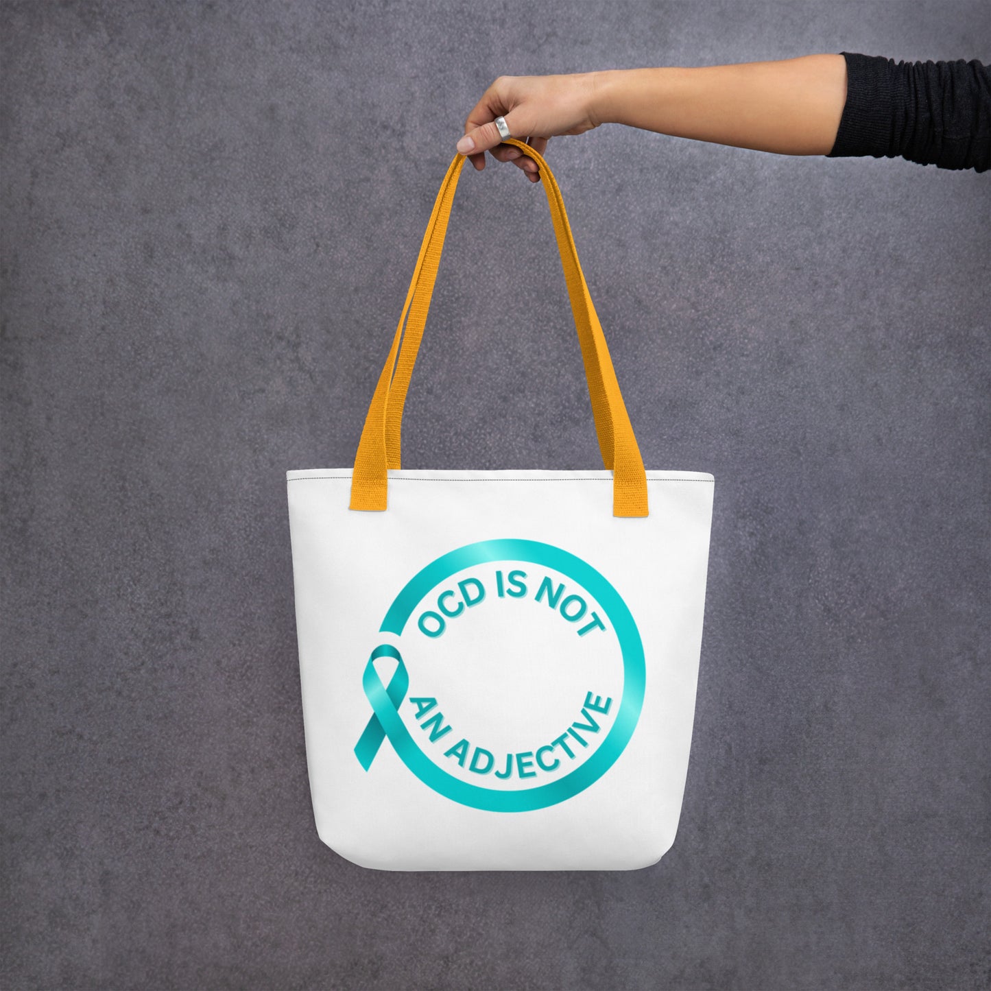 A hand holds NeuroNiche Design’s "OCD Is Not An Adjective" white tote bag with yellow straps, featuring a teal awareness ribbon and circular design with the phrase in teal on a plain gray background.
