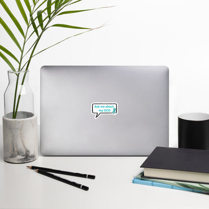 A silver laptop featuring the "Ask Me About My OCD" vinyl sticker from NeuroNiche Design sits on a white desk next to two black pens, a stack of books, and a glass vase with green leaves.