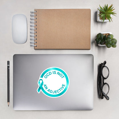 A laptop featuring NeuroNiche Design’s “OCD is Not An Adjective” vinyl sticker rests on a desk, neatly arranged with a pencil, glasses, mouse, notebook, and two potted plants.