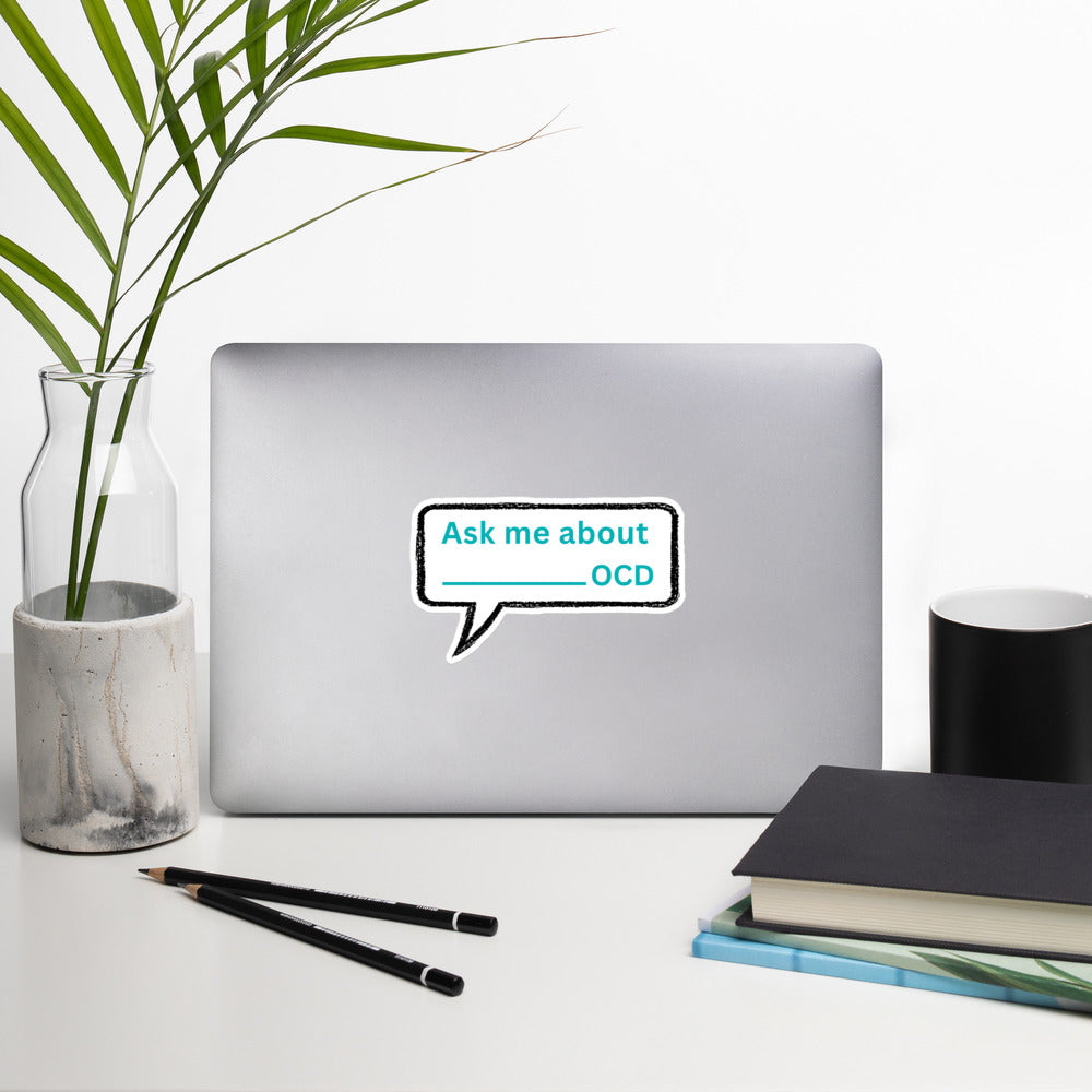 A silver laptop displays NeuroNiche Design’s "Ask Me About My OCD" high-opacity vinyl sticker on a white desk with a plant in a glass vase, two pens, black notebook, and white cup—enjoy easy, bubble-free application.