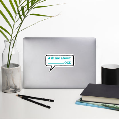 A silver laptop displays NeuroNiche Design’s "Ask Me About My OCD" high-opacity vinyl sticker on a white desk with a plant in a glass vase, two pens, black notebook, and white cup—enjoy easy, bubble-free application.