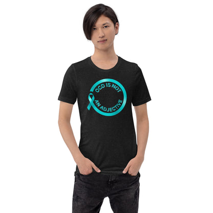 A person with medium-length dark hair stands with hands in pockets, wearing NeuroNiche Design’s OCD Is Not An Adjective T-shirt featuring a teal ribbon and “OCD IS NOT AN ADJECTIVE” inside a teal circle.