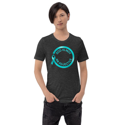 A person with straight dark hair, hands in pockets, models the NeuroNiche Design "OCD Is Not An Adjective" t-shirt—a dark combed cotton tee with a teal awareness ribbon and bold text on the front.