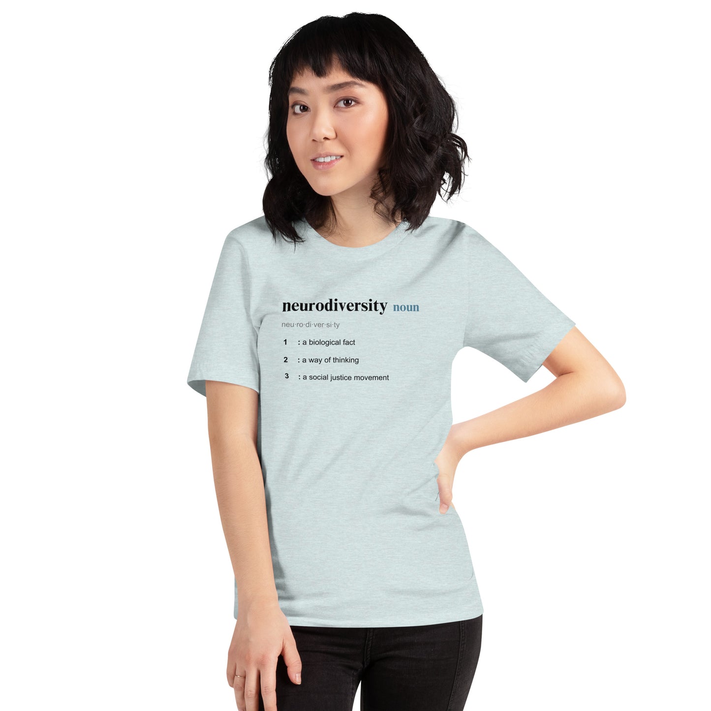 A young woman with shoulder-length black hair wears NeuroNiche Design's "Defining Neurodiversity" tee, a comfy light gray 100% cotton shirt featuring the phrase "neurodiversity noun" and three definitions. She stands, smiling slightly with one hand on her hip.